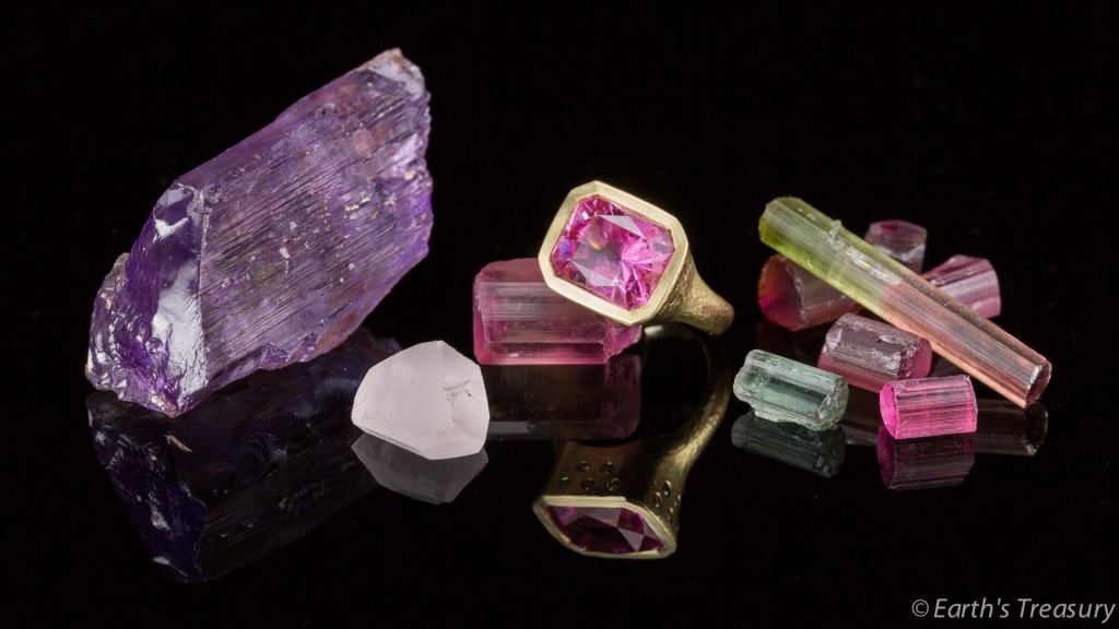 A collection of stunning pieces from the California Gems family photo content, featuring a large purple rough crystal, small multicolored crystals, a pink faceted stone ring, and various cut and rough gems all displayed on a black reflective surface, evoke the vibrant spirit of Southern California.