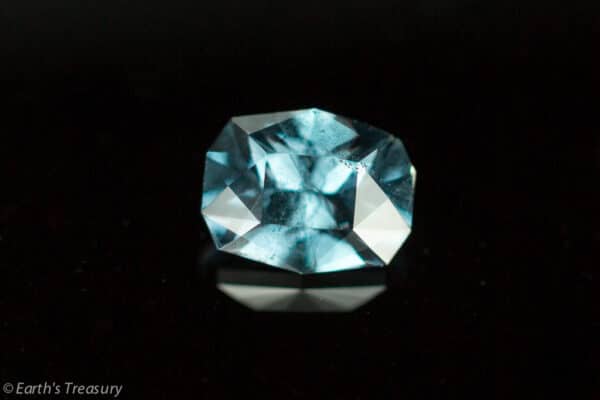 _56A8801-1024 The 0.88-Carat Blue-Violet Color Change Garnet, expertly faceted with multiple cuts, reflects light beautifully and showcases a captivating blend of darker and lighter blue shades. Positioned against a solid black background, its transparent and sparkling qualities are accentuated. The bottom left corner credits Earth’s Treasury for this remarkable gemstone.