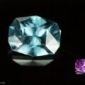 An image showing two gemstones: a substantial, aquamarine stone on the left, and a smaller, blue-violet color change garnet on the right. Both are placed on a reflective black surface, emphasizing their radiant facets and vibrant colors. Text "© Earth's Treasury" is visible in the lower-left corner.