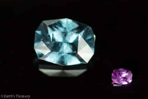 Blue-Violet Color Change Garnet An image showing two gemstones: a substantial, aquamarine stone on the left, and a smaller, blue-violet color change garnet on the right. Both are placed on a reflective black surface, emphasizing their radiant facets and vibrant colors. Text "© Earth's Treasury" is visible in the lower-left corner.