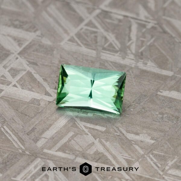 A close-up image of the 3.90-Carat Neon Blue-Green Indicolite Tourmaline, a light green rectangular-cut gemstone, placed on a textured, metallic surface. The gemstone exhibits a star-like pattern in its facets, with the Earth’s Treasury logo visible at the bottom of the image.