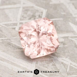 A 1.84-carat Pink-Peach Pastel Pyrope Garnet with a cushion cut rests on a textured gray surface, featuring the words "EARTH'S TREASURY" and a hexagonal logo beneath it.