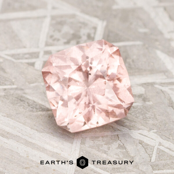 A 1.84-carat Pink-Peach Pastel Pyrope Garnet with a cushion cut rests on a textured gray surface, featuring the words "EARTH'S TREASURY" and a hexagonal logo beneath it.