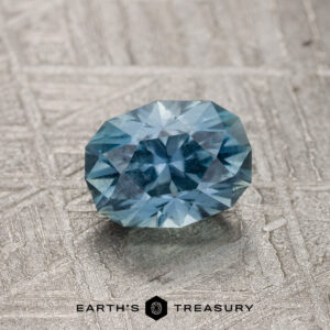 A 1.33-Carat Gray-Aqua Montana Sapphire rests on a textured metallic surface, with the Earth's Treasury logo and text displayed below.