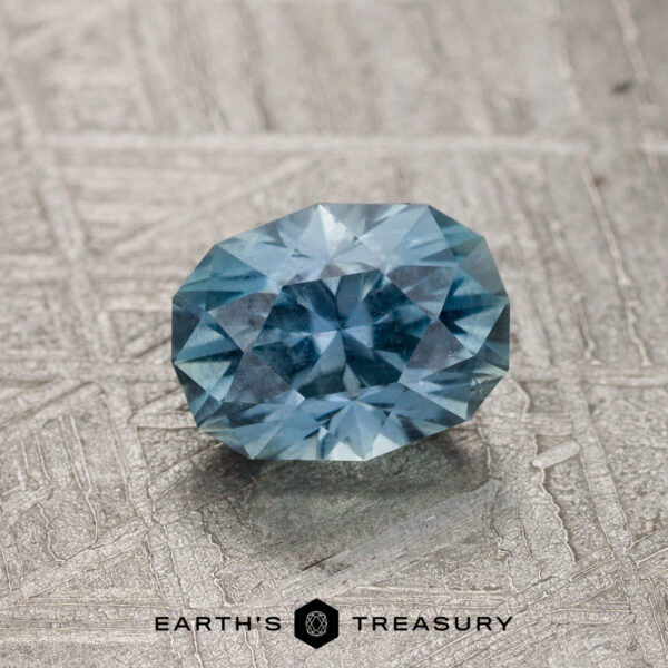 1.33-Carat Gray-Aqua Montana Sapphire A 1.33-Carat Gray-Aqua Montana Sapphire rests on a textured metallic surface, with the Earth's Treasury logo and text displayed below.