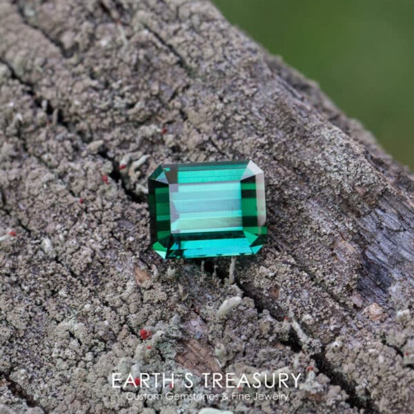 Shaded Daylight: 12.09-Carat Blue-Green Afghan Tourmaline A rectangular 12.09-carat blue-green Afghan tourmaline with an emerald cut is displayed on a rough textured log. The text at the bottom reads, "Earth's Treasury - Custom Gemstones & Fine Jewelry.