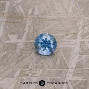 A 1.10-carat bright blue Umba sapphire is set against a textured gray background. The gemstone is intricately faceted, displaying a vibrant mix of light and dark blue hues. At the bottom of the image, the text reads "Earth's Treasury" with a geometric logo beside it.