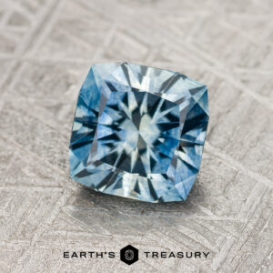 A 1.63-carat medium blue Montana Sapphire (heated) with a square cut rests on a textured surface, with "EARTH'S TREASURY" shown below.