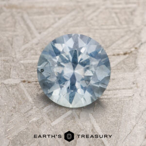 A 1.13-carat light aqua blue Montana sapphire displayed on a textured background. The gemstone, featuring a light blue hue, catches the light, revealing its facets. Below, text reads "EARTH'S TREASURY" with a logo icon between the words.