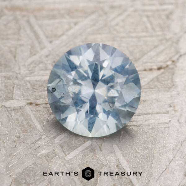 1.13-Carat Light Aqua Blue Montana Sapphire A 1.13-carat light aqua blue Montana sapphire displayed on a textured background. The gemstone, featuring a light blue hue, catches the light, revealing its facets. Below, text reads "EARTH'S TREASURY" with a logo icon between the words.