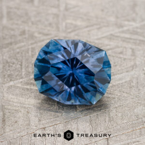 A 1.45-carat Royal Blue Montana Sapphire (heated) is showcased on a textured gray surface, with the "Earth's Treasury" logo and text at the bottom of the image.