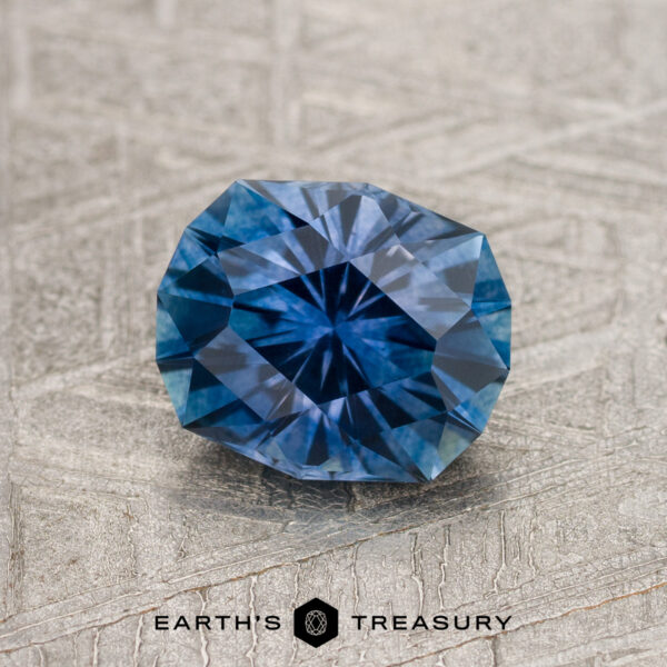1.45-Carat Royal Blue Montana Sapphire (Heated) A 1.45-carat Royal Blue Montana Sapphire (heated) is showcased on a textured gray surface, with the "Earth's Treasury" logo and text at the bottom of the image.