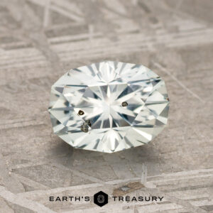 A 1.58-carat near colorless Montana Sapphire with visible inclusions, oval-cut, rests on a textured surface. The Earth's Treasury logo appears below.