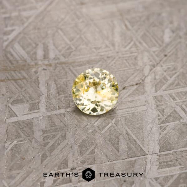 ET1273 A 1.12-Carat Yellow Ceylon Sapphire (Heated), featuring a round cut and pale yellow hue, rests on a textured gray surface. The background is adorned with intricate, intersecting line patterns. At the bottom of the image, the logo and text "Earth's Treasury" are displayed.