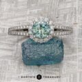 The "Charlotte" ring in platinum, featuring a 1.31-carat Montana sapphire and halo setting, is displayed on a rough blue gemstone atop a patterned surface.