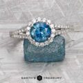 The "Charlotte" ring in 14K white gold features a 1.71-carat Montana sapphire, diamond halo, and diamond-accented band, shown on a rough blue crystal with the Earth’s Treasury logo below.