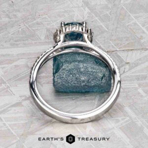 The "Charlotte" ring in 14K white gold with a 1.71-carat Montana sapphire is displayed upright against a rough blue mineral on a textured gray surface, with the Earth's Treasury logo at the bottom.