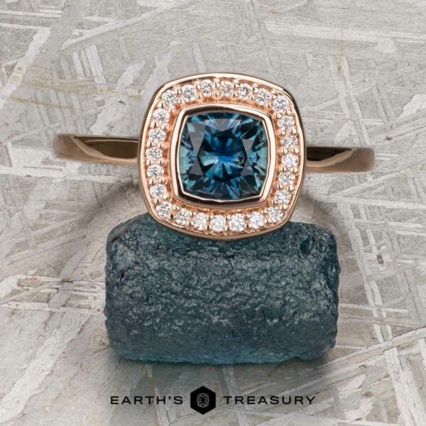 The "Grace" ring in 14K rose gold with 0.90-carat Montana sapphire The "Grace" ring in 14K rose gold features a 0.90-carat cushion-cut Montana sapphire, encircled by a halo of diamonds and displayed on a rough blue stone against a textured gray background.