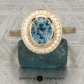The “Grace” ring in 14K yellow gold showcases a 1.74-carat Montana sapphire with a bezel-set halo of diamonds, elegantly displayed on a rough blue stone above the "Earth's Treasury" logo.