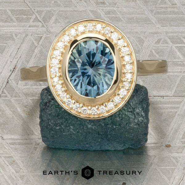 The “Grace” ring in 14K yellow gold with 1.74-carat Montana sapphire The “Grace” ring in 14K yellow gold showcases a 1.74-carat Montana sapphire with a bezel-set halo of diamonds, elegantly displayed on a rough blue stone above the "Earth's Treasury" logo.