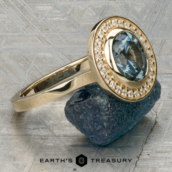 The “Grace” ring in 14K yellow gold with 1.74-carat Montana sapphire The “Grace” ring in 14K yellow gold showcases a 1.74-carat Montana sapphire with a bezel-set halo of diamonds, elegantly displayed on a rough blue stone above the "Earth's Treasury" logo.