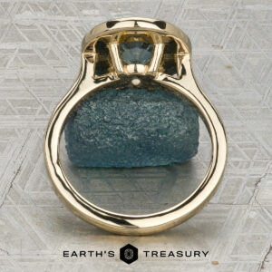 The “Grace” ring in 14K yellow gold showcases a 1.74-carat Montana sapphire with a bezel-set halo of diamonds, elegantly displayed on a rough blue stone above the "Earth's Treasury" logo.