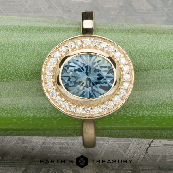 The “Grace” ring in 14K yellow gold with 1.74-carat Montana sapphire The “Grace” ring in 14K yellow gold showcases a 1.74-carat Montana sapphire with a bezel-set halo of diamonds, elegantly displayed on a rough blue stone above the "Earth's Treasury" logo.