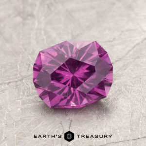 A faceted ET1123 gemstone in purple rests on a textured gray surface.
