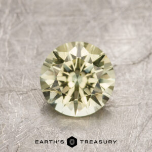 A 1.36-carat greenish-yellow Montana Sapphire (heated) is shown on a textured surface, radiating brilliance above the Earth’s Treasury logo and name at the bottom.