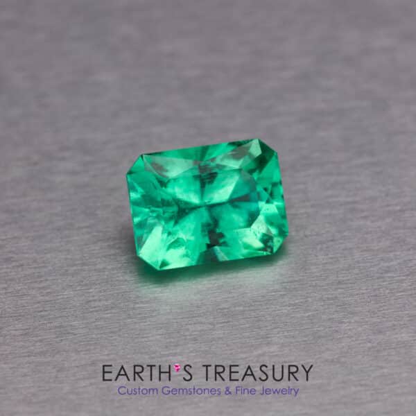 _54A2446-ET-1200 A vibrant 1.48-carat Ethiopian emerald, expertly cut in an emerald shape, is placed on a smooth, metallic surface. Below the gemstone, the text reads "EARTH'S TREASURY - Custom Gemstones & Fine Jewelry." The gemstone features clean, sharp facets and a brilliant, rich green color.