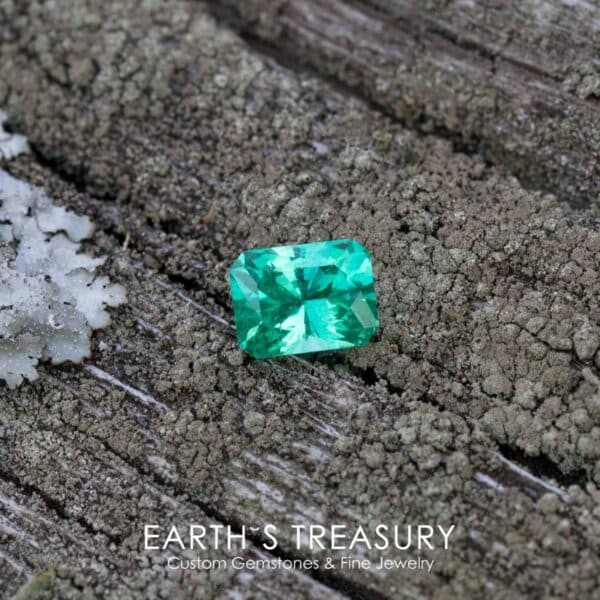 _B3A3423-ET-1200 A vibrant 1.48-carat Ethiopian emerald, rectangular in shape, rests on a weathered, textured wooden surface. Moss and lichen surround the gemstone. Text at the bottom of the image reads "EARTH'S TREASURY Custom Gemstones & Fine Jewelry.