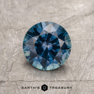A 1.62-carat deep blue heated Montana Sapphire, round and faceted, is shown on a textured gray surface with the "Earth's Treasury" logo and text at the bottom.