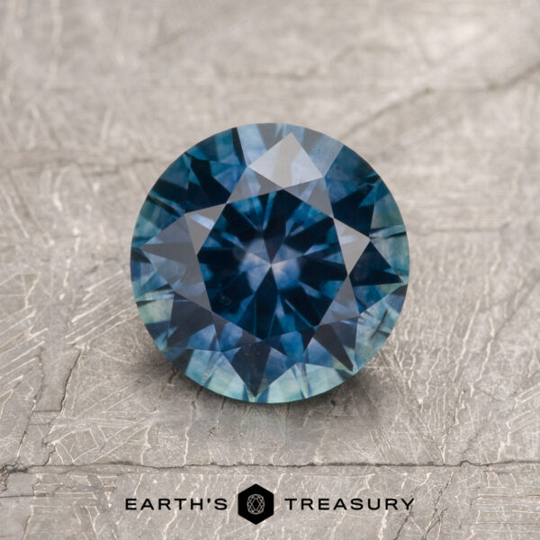 1.62-Carat Deep Blue Montana Sapphire (Heated) A 1.62-carat deep blue heated Montana Sapphire, round and faceted, is shown on a textured gray surface with the "Earth's Treasury" logo and text at the bottom.