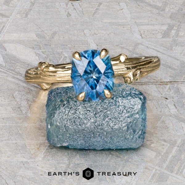 The "Twyg" ring in 18K yellow gold with 1.53-carat Montana sapphire The "Twyg" ring in 18K yellow gold, featuring a 1.53-carat Montana sapphire, rests on a rough blue stone atop a textured gray surface. "Earth's Treasury" appears below, reflecting the natural elegance of Twyg.
