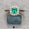 The "Anne" ring in 14K yellow gold with a 0.71-carat emerald and diamond halo rests on a rough blue gemstone, with the "Earth's Treasury" logo below.