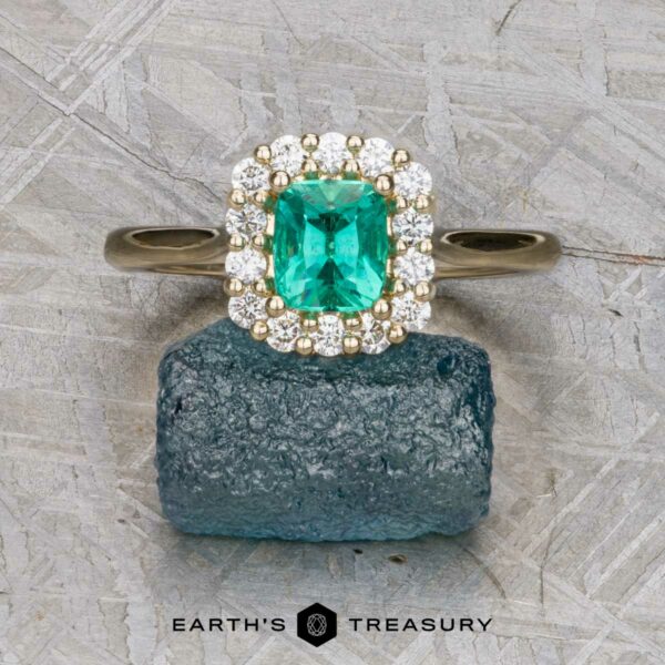 The "Anne" ring in 14K yellow gold with 0.71-carat emerald The "Anne" ring in 14K yellow gold with a 0.71-carat emerald and diamond halo rests on a rough blue gemstone, with the "Earth's Treasury" logo below.