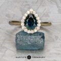 The "Anne" ring in 14k yellow gold, featuring a 1.44-carat Australian sapphire and diamond halo, is shown atop a rough blue gemstone on a textured background with the "Earth's Treasury" logo below.