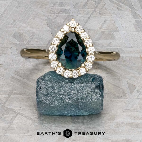 The "Anne" ring in 14k yellow gold with 1.44-carat Australian sapphire The "Anne" ring in 14k yellow gold, featuring a 1.44-carat Australian sapphire and diamond halo, is shown atop a rough blue gemstone on a textured background with the "Earth's Treasury" logo below.
