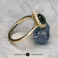 The "Anne" ring in 14k yellow gold with a 1.44-carat Australian sapphire rests against a rough blue stone on a textured gray surface, with the Earth's Treasury logo at the bottom.