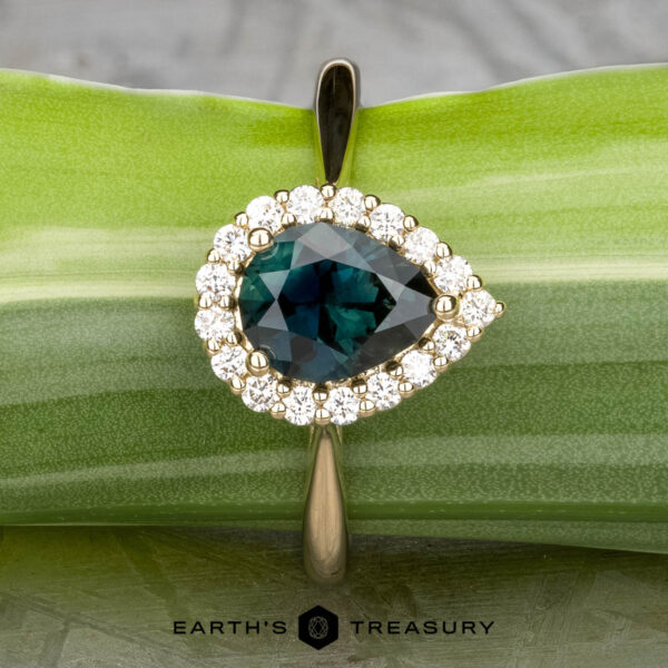 The "Anne" ring in 14k yellow gold with 1.44-carat Australian sapphire The "Anne" ring in 14k yellow gold features a 1.44-carat Australian sapphire, pear-shaped and surrounded by diamonds, displayed on a green leaf with "EARTH'S TREASURY" below.
