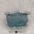 The Petite Pave Contour Band in platinum with Montana sapphire rests on a rough blue-green stone, set against a textured gray background. The "Earth's Treasury" logo appears at the bottom.