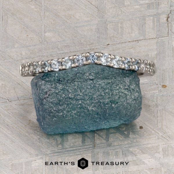 The Petite Pave Contour Band in platinum with Montana sapphire The Petite Pave Contour Band in platinum with Montana sapphire rests on a rough blue-green stone, set against a textured gray background. The "Earth's Treasury" logo appears at the bottom.