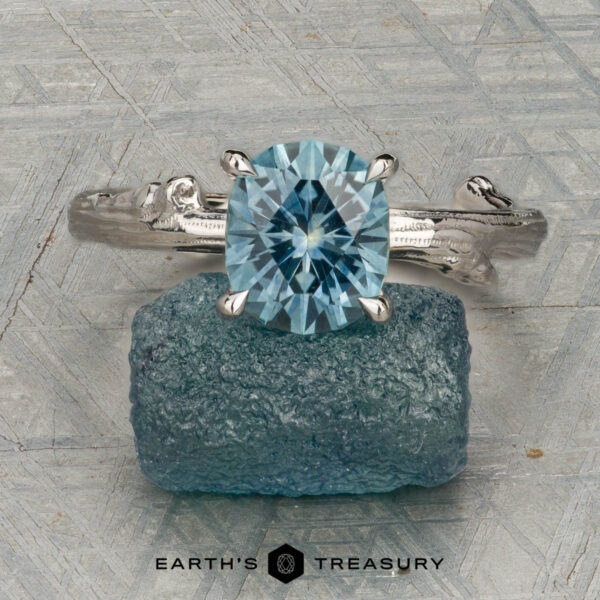 The "Twyg" ring in 14K white gold with 2.05-carat Montana sapphire The "Twyg" ring in 14K white gold, featuring a 2.05-carat Montana sapphire, is displayed atop a rough blue stone with the text "Earth's Treasury" below.