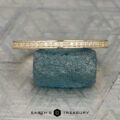 The Straight "Grace" band in 14K yellow gold with small diamonds rests on a rough blue-green stone atop a patterned gray surface. "EARTH'S TREASURY" appears at the bottom.