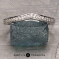 A platinum “Faye” band encircles a rough, dark green stone, displayed on a textured gray surface with the Earth’s Treasury logo at the bottom.