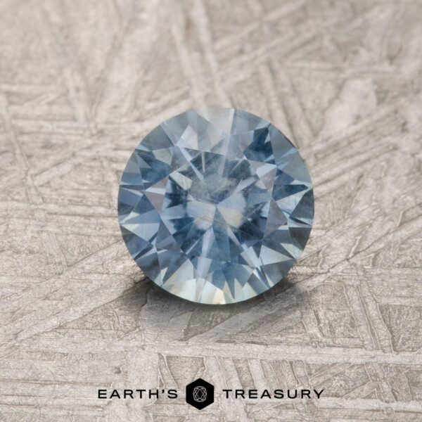 1.01-Carat Light Aqua Blue Montana Sapphire A 1.01-carat light aqua blue Montana sapphire, round-cut and radiant, rests on a textured gray surface with "EARTH'S TREASURY" written below.