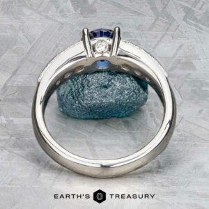 The Cascade ring in platinum with a 1.24-carat Ceylon sapphire is displayed upright against a blue rock on textured gray, with the "Earth's Treasury" logo at the bottom.