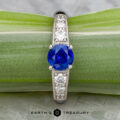 The "Cascade" ring in platinum features a 1.24-carat Ceylon sapphire at the center, accented by smaller clear stones on each side and elegantly displayed on a green plant stem.