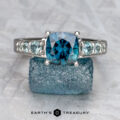 The "Cascade" ring in 14K white gold features a 2.08-carat Australian sapphire, flanked by smaller stones, shown on a rough blue crystal with the "Earth's Treasury" logo below.