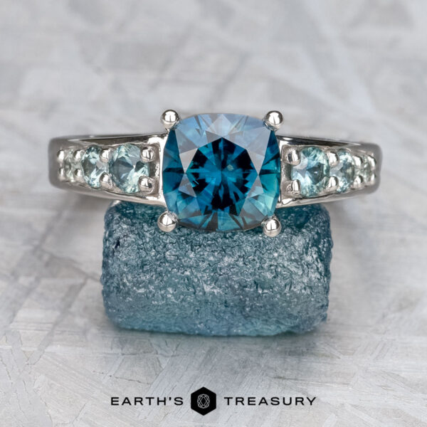 The "Cascade" ring in 14K white gold with 2.08-carat Australian sapphire The "Cascade" ring in 14K white gold features a 2.08-carat Australian sapphire, flanked by smaller stones, shown on a rough blue crystal with the "Earth's Treasury" logo below.